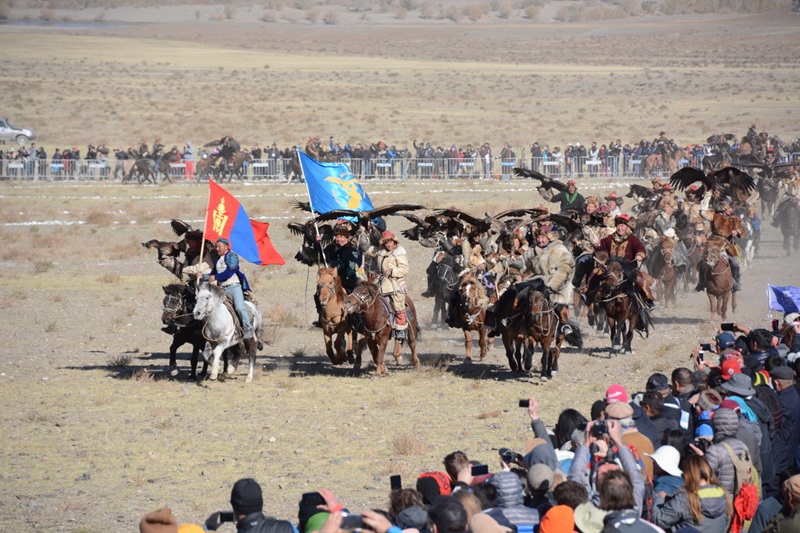 A large group of traditional Mongolian eagle hunters on horseback carrying flags and golden eagles during the festival opening parade.