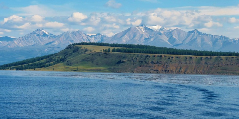A wide view of the crystal blue waters of Lake Kh&ouml;vsg&ouml;l with a forested shoreline and snow-capped Sayan Mountains in the background.