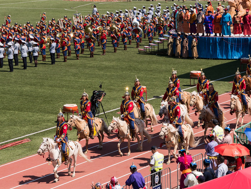 Mongolian State Honor Guard in traditional red and blue uniforms riding white horses during the Naadam Festival opening ceremony in a stadium.