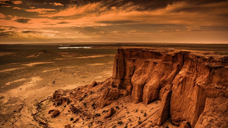 High-angle view of the colorful sedimentary rock formations at Tsagaan Suvarga (White Stupa) in Mongolia under a vibrant orange and pink sunset sky.